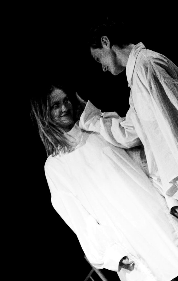 A figure in white seated, smiling, with a second figure standing close
