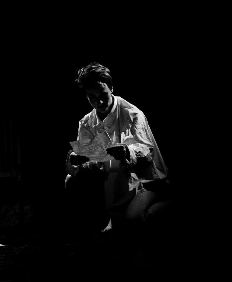 A kneeling figure in deep shadow, examining a fragment of paper