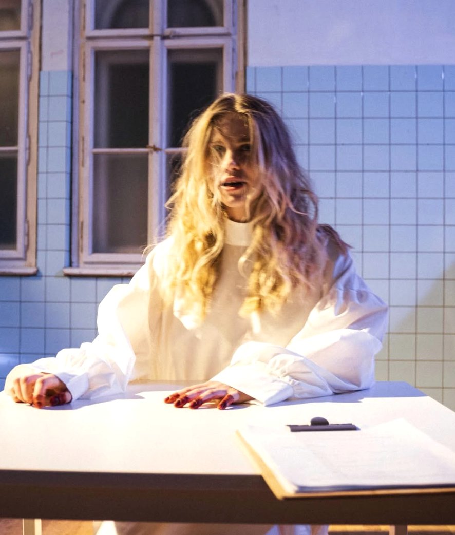A woman in white at a table, blood on her hands, a stark gaze